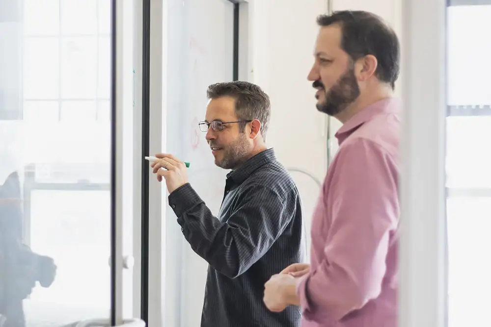 Two men standing infront of a whiteboard while one of them holds a marker in his hand