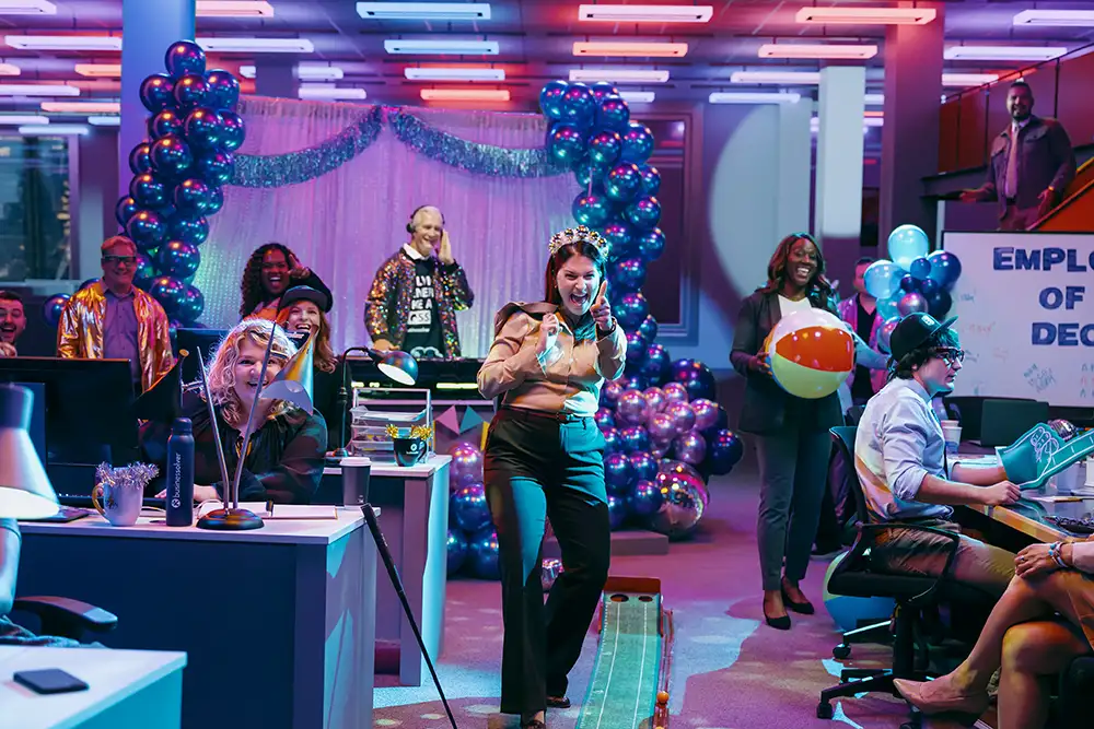 A businesswoman wearing a crown laughing with an office party happening behind her